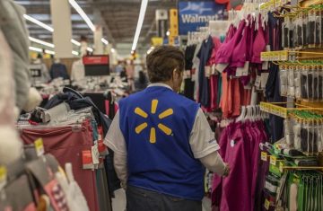 A retail employee wearing a blue vest with a Walmart logo walking past crowded store clothing racks while using a Walmart handheld radio.