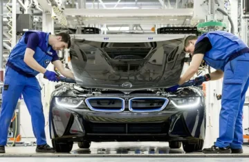 Two automotive assembly line workers in blue uniforms working under the hood of a black BMW vehicle