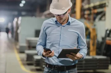 Industrial Engineer Using Mobile Devices in Smart Manufacturing Engineer in a factory using a smartphone and tablet while wearing a hard hat.