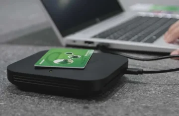 A black RFID reader with a green RFID card placed on top, connected to a laptop via a USB cable.