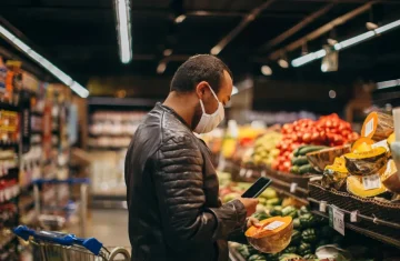 Man wearing mask checking smartphone while shopping for fresh vegetables in supermarket.