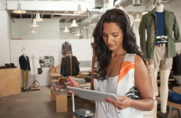 Retail manager checks inventory on a tablet inside a clothing store with apparel displays and mannequins.