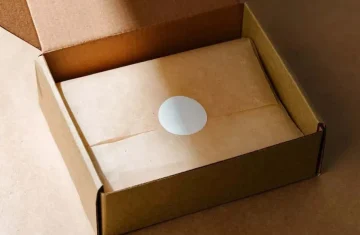 A small parcel wrapped in brown paper and sealed with a white circular sticker, placed inside a cardboard box