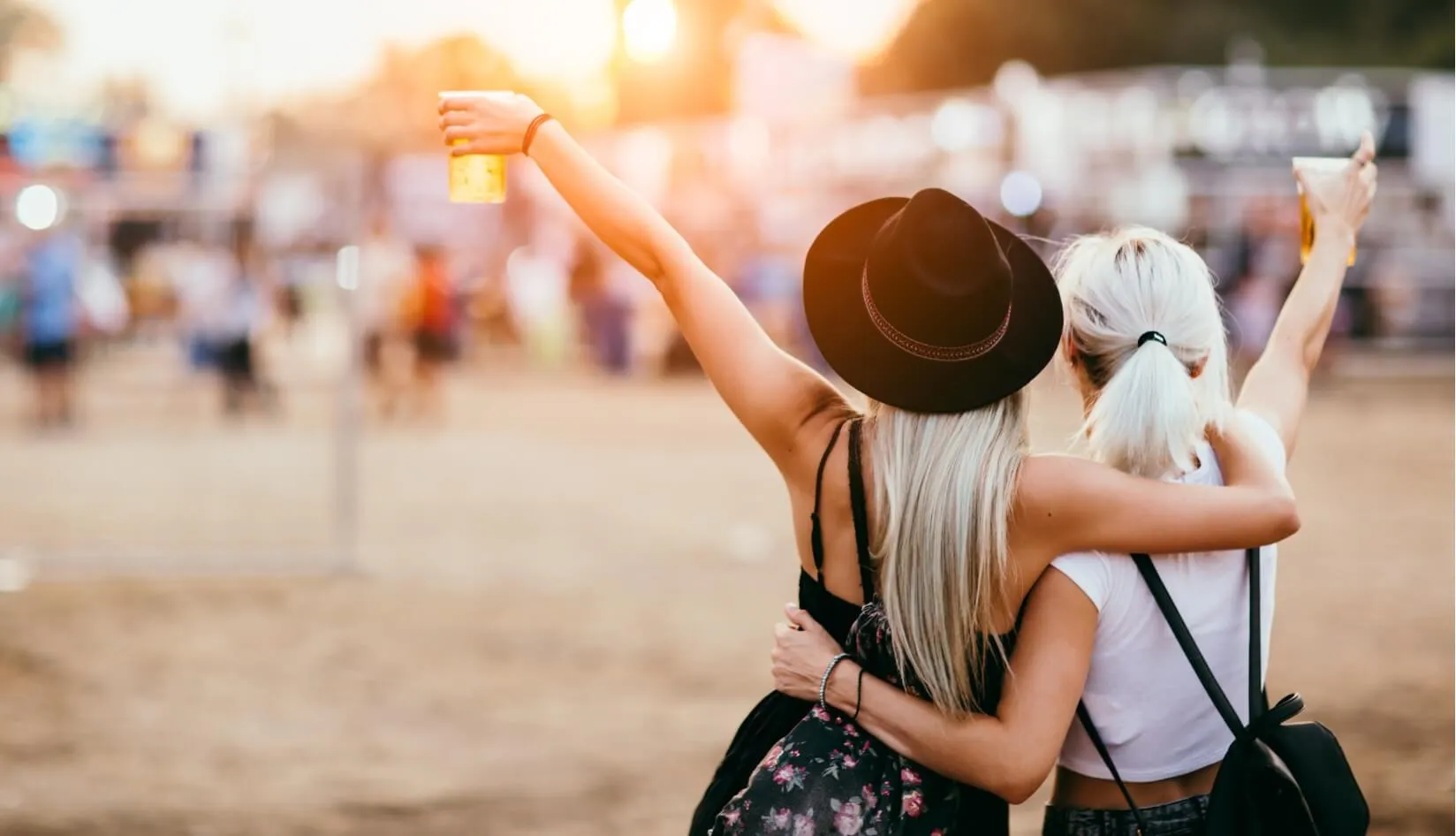 Enjoying the Festival Together Two women at a music festival raising their glasses to celebrate the event, with a crowd in the background.