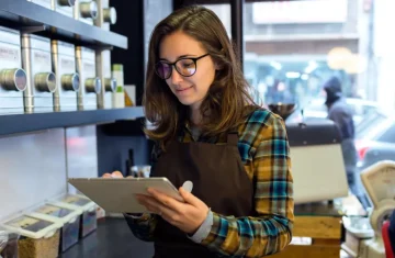Cafe staff member wearing an apron uses a tablet near coffee containers and equipment