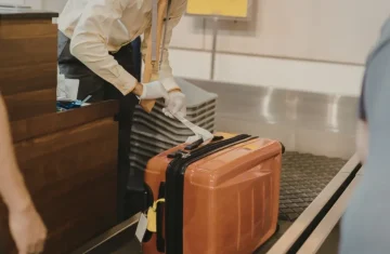 Airport Check-in and Baggage Handling An airline check-in agent in a yellow shirt and white gloves applies a baggage tag to an orange suitcase on a conveyor belt.