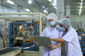 Two female factory workers in hairnets, face masks, and lab coats looking at a laptop computer in front of a production line.