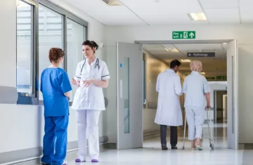 A doctor and a nurse talking in a bright hospital corridor, while another doctor assists an elderly patient in the background.