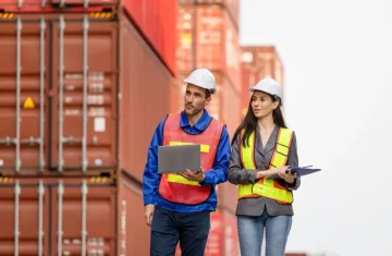 A male and a female logistics manager in hard hats and safety vests walking and talking in a container yard, with one holding a laptop.