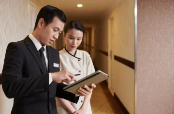 A male hotel manager in a suit showing a tablet to a female housekeeper in uniform in a hotel hallway.