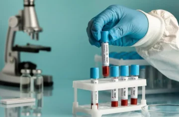 A gloved hand in a lab coat holding a blood sample test tube labeled "COVID-19 TEST" over a rack, with a microscope in the background.