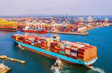 An aerial view of a large container ship loaded with colorful containers being guided by tugboats into a busy commercial port.