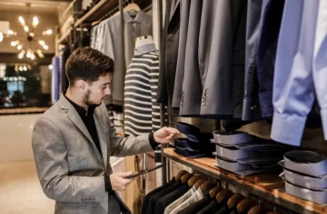 Smart Retail: Inventory Management and Product Information A male shopper in a suit jacket examines folded shirts on a shelf in a modern, well-lit clothing boutique.