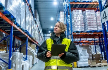 Real-Time Inventory Tracking in Cold Storage and Warehouses A female warehouse worker in a high-visibility vest and winter jacket looks up at tall shelving units in a cold storage or large warehouse.