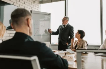 A senior male executive in a suit giving a presentation with a line graph on a whiteboard to his team in a conference room.