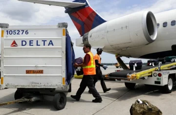Delta ground crew workers in orange safety vests loading luggage from a baggage cart onto a conveyor belt leading into an airplane.