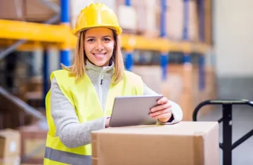 Beyond the Blurred Barcode Why RFID is the Ultimate Solution for Flawless Data Capture Smiling warehouse worker in a yellow hard hat and high-visibility vest holding a tablet beside a cardboard box in a warehouse aisle.