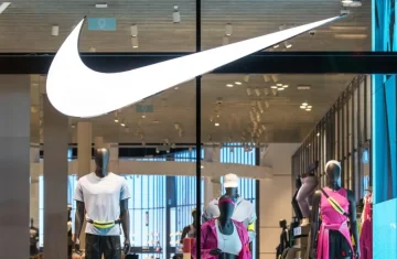 Illuminated Nike "swoosh" logo above the entrance of a Nike retail store, with mannequins displaying sportswear inside.