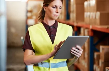 A woman in a yellow safety vest using a tablet in a warehouse with shelves of boxes in the background.