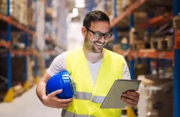 The Visible & The Invisible Unlocking Value with Custom RFID Asset Stickers A smiling warehouse worker wearing a yellow safety vest and glasses, holding a tablet and a blue hard hat in a warehouse.