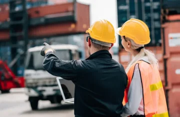 Two logistics workers wearing yellow hard hats review a laptop while pointing toward stacked shipping containers in a busy freight yard.