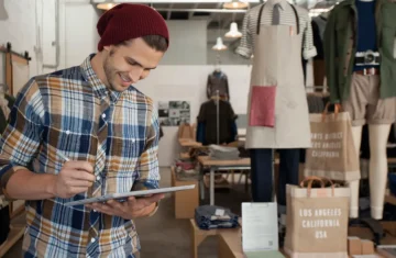 A man writing on a tablet while standing in a clothing store with clothes displayed in the background