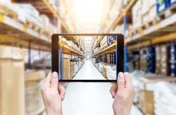 RFID Asset Tracking News How GS1’s New TDS 2.3 Standard Gives RFID Tags Their Own Web Address Hands holding a tablet in a warehouse aisle, showing shelves, boxes, and inventory storage in a logistics setting.