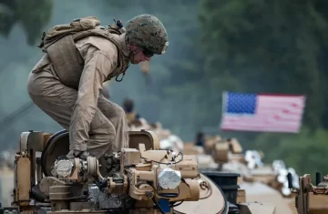 Soldier climbing on a military vehicle during field operations, with other vehicles and an American flag in the background.