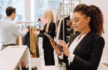 Retail staff using a tablet in an apparel store, team members organizing clothing displays in the background.