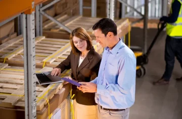 RFID Label Tooling Charge Warehouse staff reviewing pallets with laptop and clipboard, representing RFID label tooling charge, mold fee, setup fee, custom label specification, inlay positioning, die cutting, material testing, and production planning.