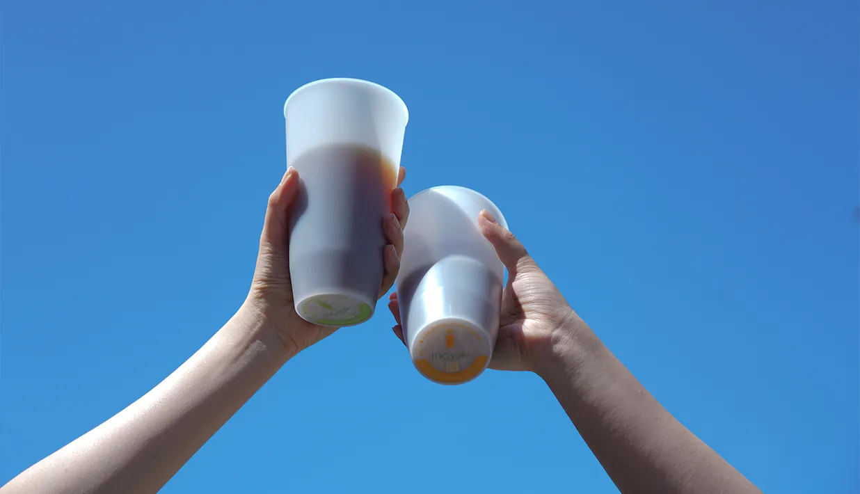 Two hands holding reusable drink cups with circular labels on their bases against a blue sky.